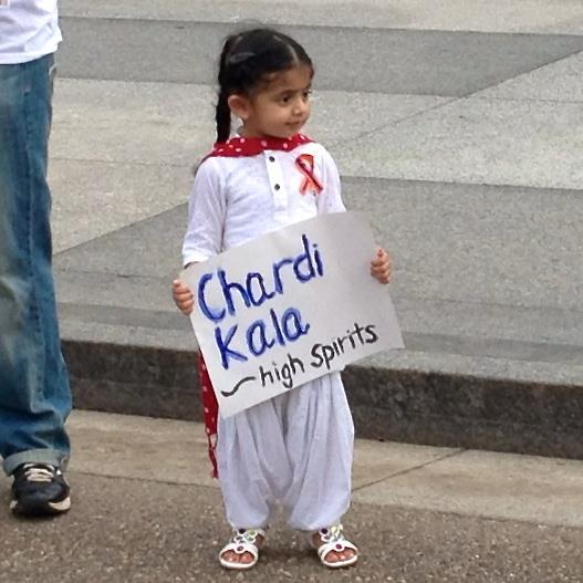A young girl holds a sign: "Chardi Kala - High Spirits". (source: Just Sikh)